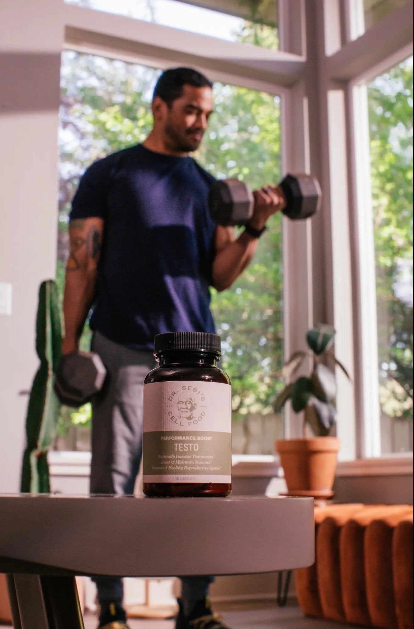 Man exercising with dumbbells beside Dr. Sebi's Performance Boost Testo supplement in bright, plant-filled home gym.