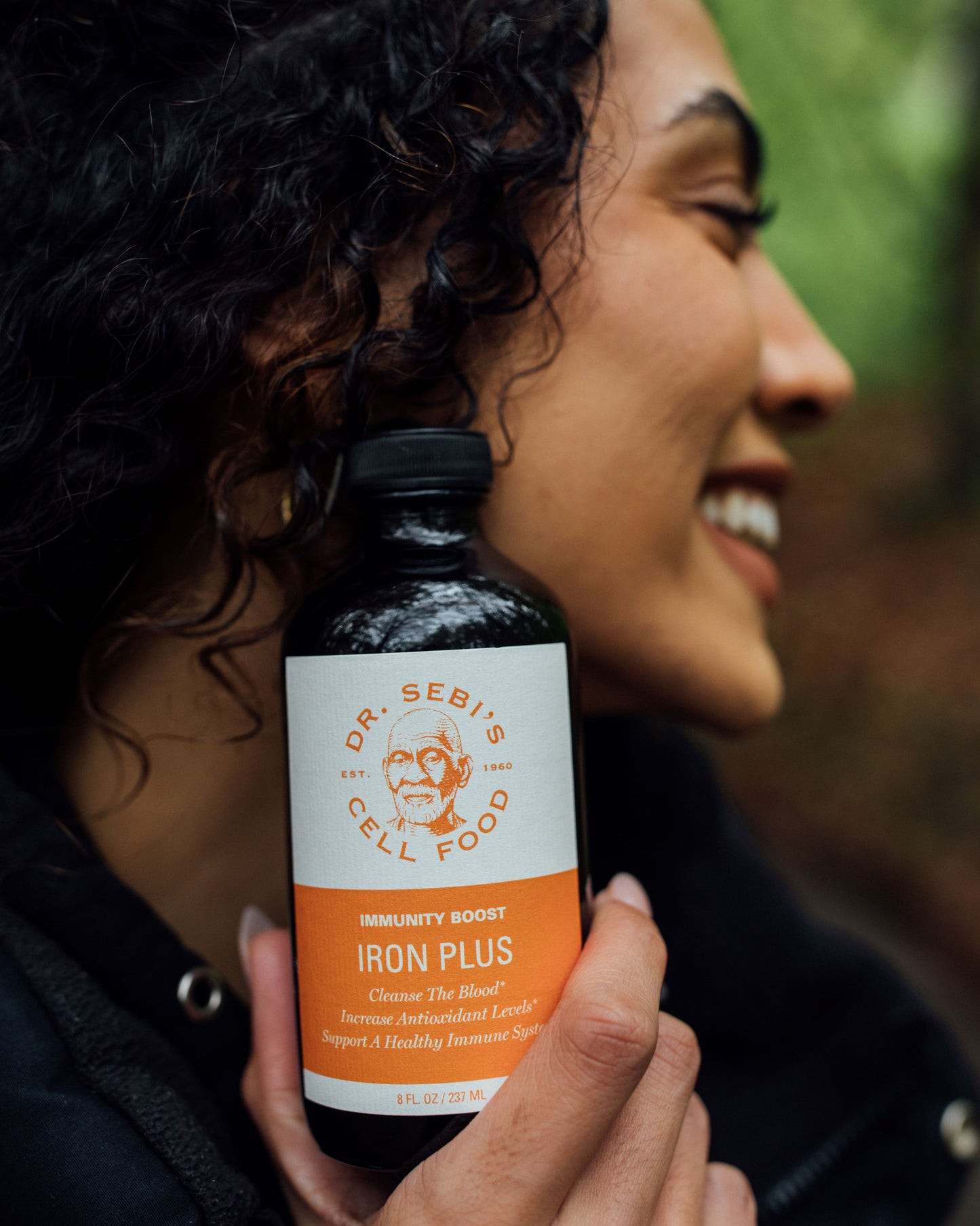 Woman holding Dr. Sebi's Iron Plus bottle outdoors, smiling with curly hair and green background.