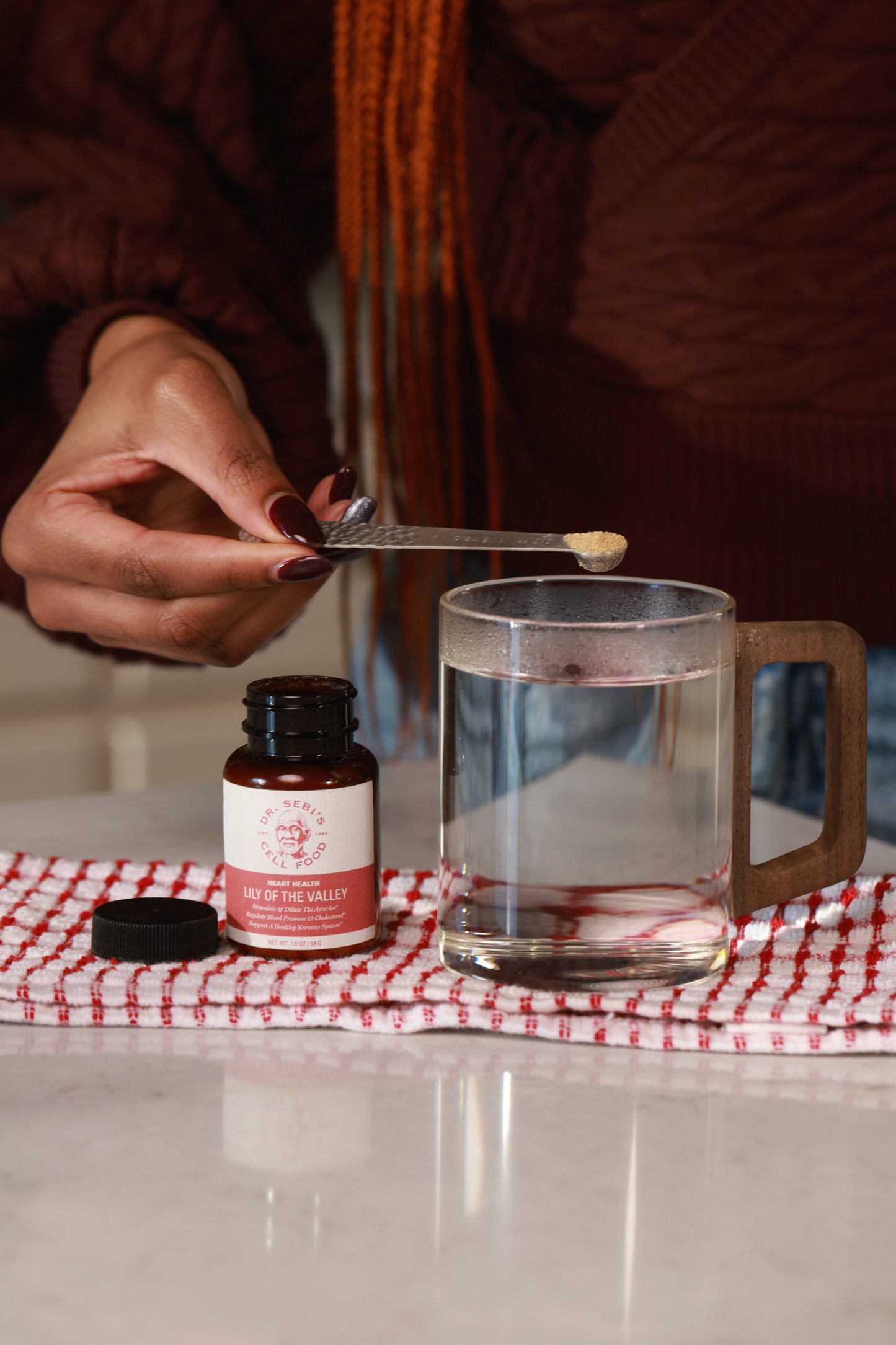 Person preparing herbal remedy with Dr. Sebi's Lily of the Valley supplement and water in wooden mug.