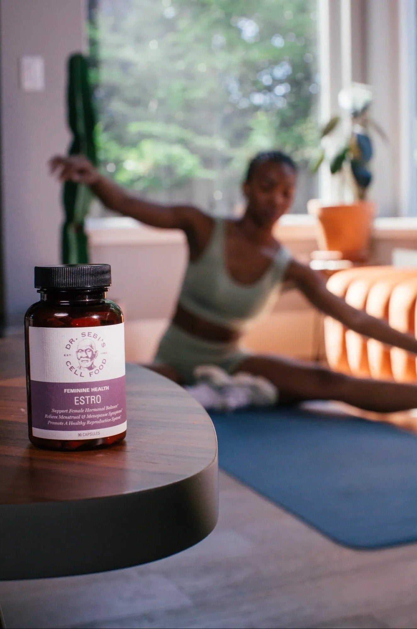Dr. Sebi's Estro supplement bottle displayed on a table with a woman doing yoga in a bright, plant-filled wellness space.