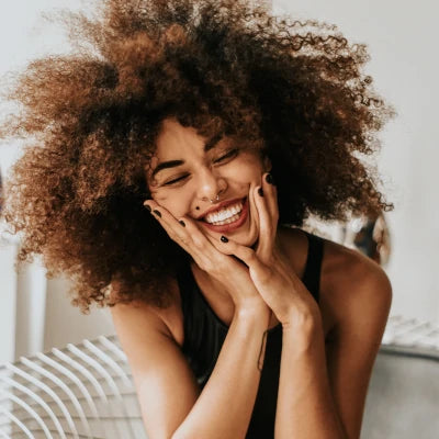 Happy woman with curly hair smiling while touching her face in a bright, wellness-focused setting.