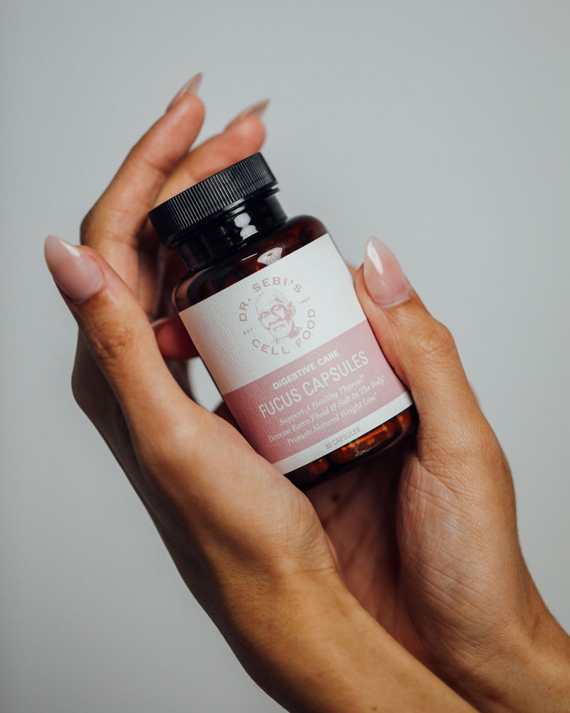 Hand holding Dr. Sebi's Cell Food Ficus Digestive Care Capsules bottle against neutral background.