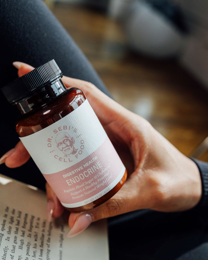 Hand holding Dr. Sebi's Cell Food Endocrine supplement bottle in amber glass with pink label outdoors.