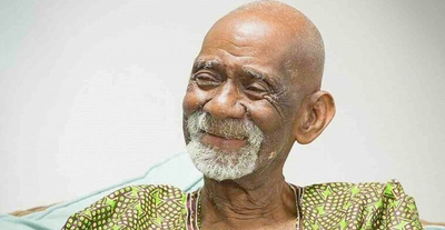 Elderly man with gray beard wearing green patterned traditional clothing, smiling warmly against a light background.