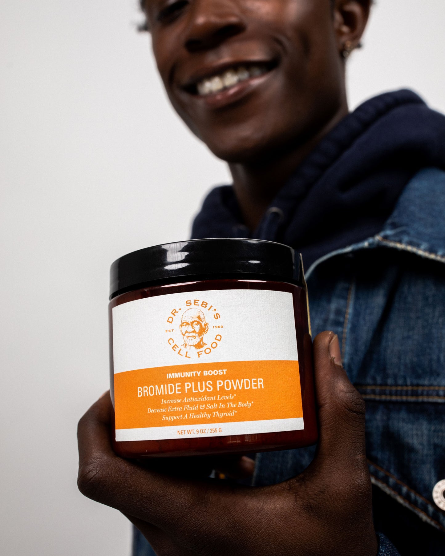 Person holding Dr. Sebi's Bromide Plus Powder jar, smiling against white background in denim jacket.