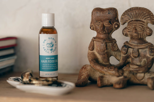 Dr. Sebi's Hair Food Oil displayed with ancient clay figurines and wellness books on wooden surface.