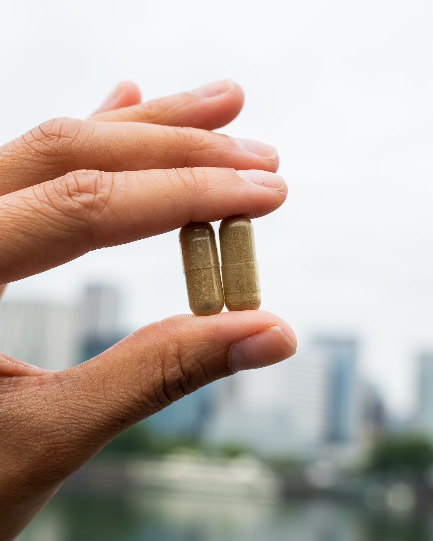 Hand holding two capsules of herbal supplement against blurred cityscape background.