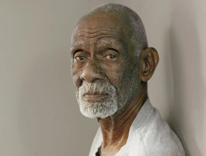Elderly man with gray beard wearing white shirt, looking thoughtfully to the side against neutral background.